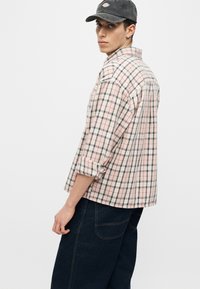 Νέος άντρας που φοράει μαύρο καπέλο, ροζ και γκρι καρό πουκάμισο με ανασηκωμένα μανίκια και σκούρα μπλε τζιν, στέκεται μπροστά σε λευκό φόντο.