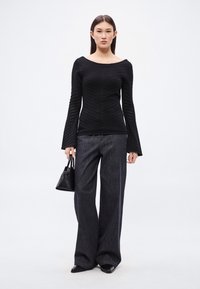 Fekete bordázott hosszú ujjú felső vállat szabadon hagyó nyakkivágással, széles szárú sötét farmernadrággal párosítva. A modell egy kis fekete kézitáskát tart.