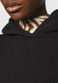 Czarna bluza z kapturem wykonana z teksturowanej tkaniny, z haftowanym logo na piersi oraz warstwowym brązowo-kremowym golfem z wzorem pod spodem.