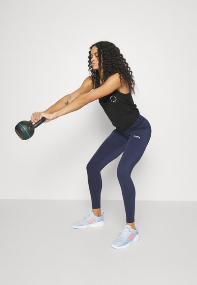 Black sleeveless top, navy leggings, and light blue athletic shoes. The model holds a kettlebell, demonstrating a workout pose.