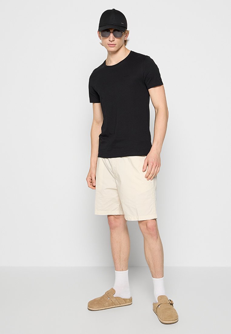 Schwarzes Kurzarm-T-Shirt, beige Shorts, braune Slippers, weiße Socken, schwarze Kappe und Sonnenbrille, vor einem neutralen Hintergrund stehend.