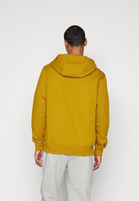 Sudadera de color mostaza con un acabado texturizado, bolsillo canguro y capucha ajustable con cordón, combinada con pantalones de chándal gris claro.