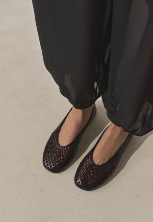 Pieds portant des ballerines en cuir tressé marron foncé associées à un pantalon noir transparent sur un sol beige clair.
