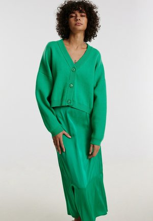 Groene cropped cardigan met knopen, geribbelde randen en lange mouwen, gecombineerd met een bijpassende groene satijnen midi-rok. Over het algemeen een gladde textuur.