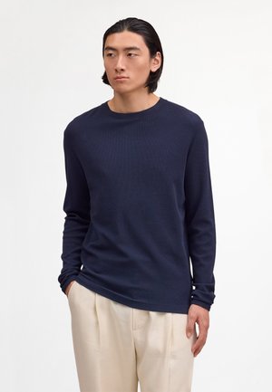 Mann med middels langt svart hår iført en marineblå langermet skjorte og lyse beige bukser, stående med hendene i lommene.