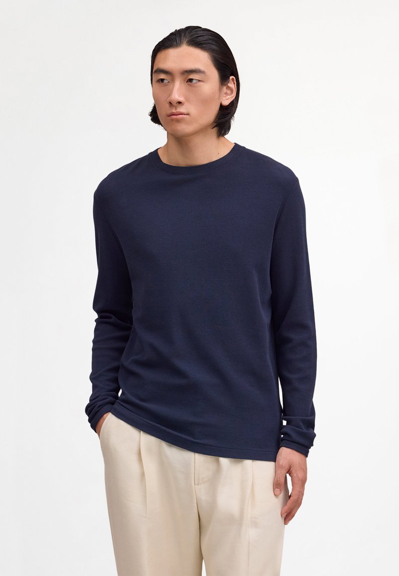 Muž so stredne dlhými čiernymi vlasmi, oblečený v tmavomodrej košeli s dlhým rukávom a svetlo béžových nohaviciach, stojaci s rukami v nohaviciach.