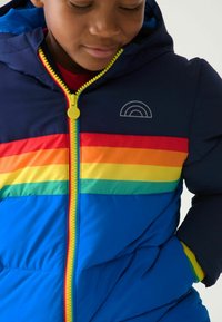 Köpeny kabát sötétkék alappal, szivárvány színű vízszintes csíkokkal és sárga cipzárokkal. Kapucnival és belső béléssel rendelkezik.