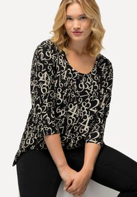 Zwarte top met een numeriek patroon in beige, met een ronde halslijn en driekwart mouwen, gestyled met donkere broeken.