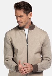 Beige gestrickte Jacke mit Reißverschluss auf der Vorderseite, kontrastierender brauner gerippter Kragen und Bündchen, mit strukturierten Ärmeln und einem glatten Körper.