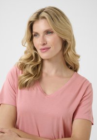 Růžové tričko s V výstřihem a krátkým rukávem, vyrobené z měkké tkaniny, nošené ženou s vlnitými blond vlasy na neutrálním pozadí.