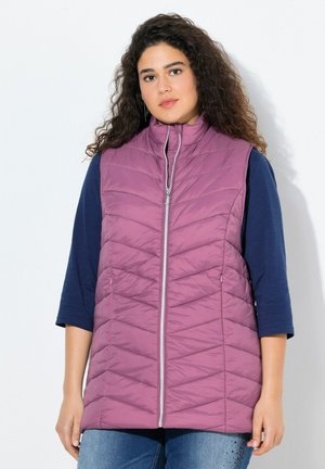 Gilet matelassé lilas avec col montant, fermeture éclair à l'avant et poches latérales, porté sur un haut bleu marine. Présente des coutures en chevrons pour ajouter de la texture.