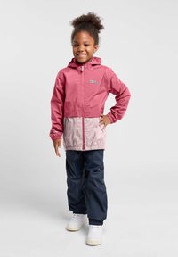 Rosa und hellgraue wasserfeste Jacke mit Kapuze, Reißverschluss vorne und elastischen Bündchen. Kombiniert mit navyblauen wasserfesten Hosen und weißen Sneakers.