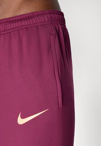 Bermudas atléticas bordeaux com um cós elástico, bolso lateral e um logotipo da Nike em laranja claro do lado esquerdo. Textura de tecido lisa e suave.