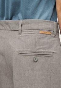 Κοντινή λήψη γκρι παντελονιών με πίσω τσέπη με κουμπί και μια μικρή ταμπελίτσα σε ταμπά χρώμα που γράφει "REPLAY SINCE 1981", φορεμένα με μπλε πουκάμισο.