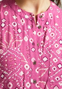 Rosa mönstrad blus med diamant- och cirkelmotiv, med knappar fram, rund halsringning och en enda bröstficka.