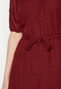 Vinröd klänning med texturerat zigzag-mönster, elastiska puffärmar och ett knytbälte i midjan. Mjukt, lätt tyg med subtil glans.