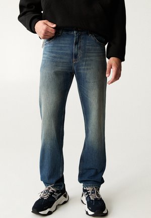 Man wearing faded blue jeans, black sweatshirt, and black and white sneakers, standing against a plain white background.