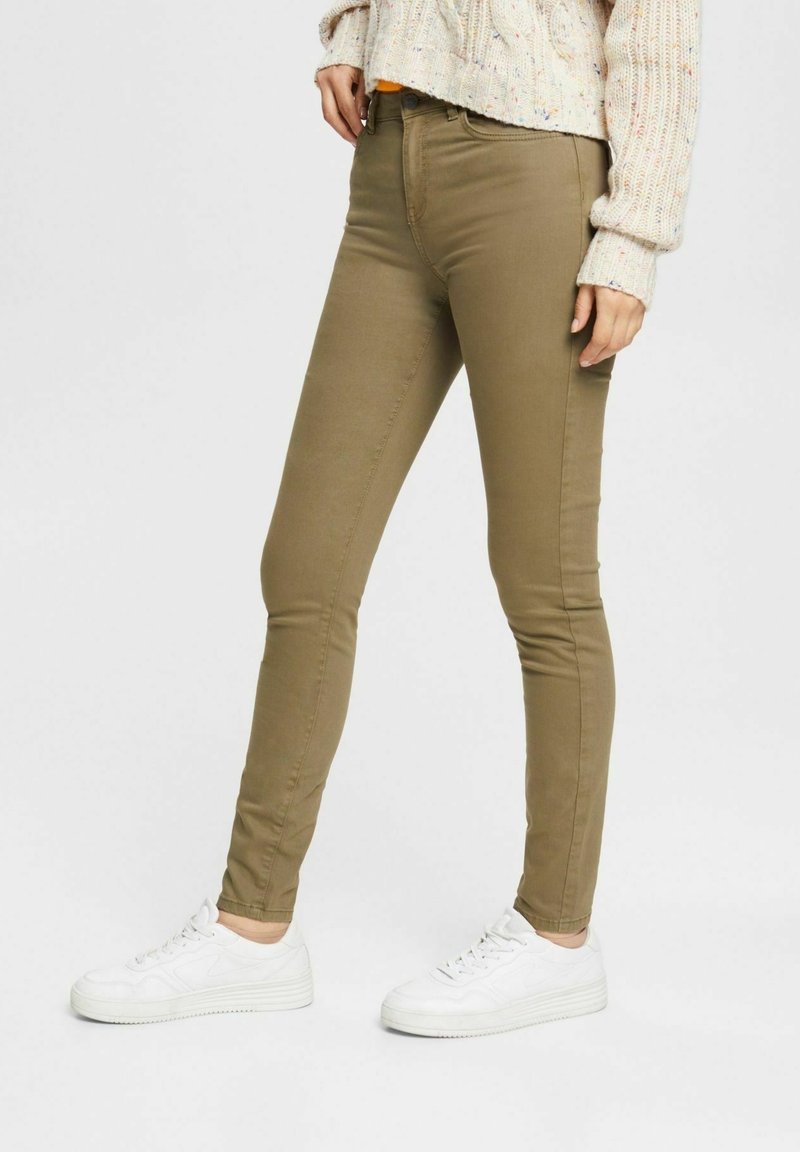 Femme portant un pantalon slim vert olive, des baskets blanches et un pull en maille crème, debout contre un fond clair uni.