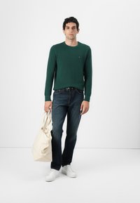 Pull en maille verte, jean foncé, baskets blanches, tenant un sac tote crème. Encolure ronde simple, design ajusté, poignets côtes.