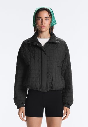 Vrouw draagt een zwarte doorgestikte jas, zwarte korte broek, wit overhemd en een groene bandana op haar hoofd, kijkt recht vooruit tegen een witte achtergrond.