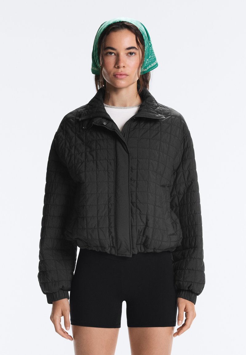 Woman wearing a black quilted jacket, black shorts, white shirt, and green bandana on her head, facing forward against a white background.