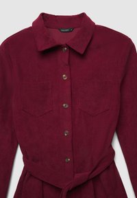 Abito in corduroy bordeaux con colletto, chiusura frontale a bottoni, due tasche sul petto e cintura annodata in vita. Presenta una texture a righe verticali.