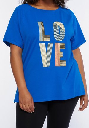 Femme portant une chemise à manches courtes bleue avec le mot "LOVE" en grosses lettres rayées, métalliques or et rose, sur le devant.