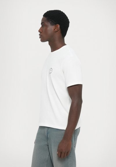 White short-sleeve t-shirt with a round neckline and a small black smiley face logo on the left chest. Worn with light gray jeans.