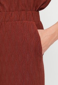 Ρούχο σε απόχρωση σκουριάς με υφή, με κυματιστό μοτίβο, ελαστική ζώνη μέσης και πλαϊνή τσέπη, που αναδεικνύει λεπτομέρεια υφασμένου υφάσματος.