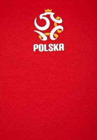 Czerwona koszulka z białym i żółtym logo oraz stylizowanymi elementami nad napisem "POLSKA" w białej czcionce, wykonana z miękkiego, teksturowanego materiału.