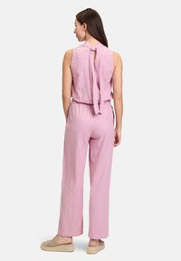 Roze mouwloze jumpsuit met een strik op de rug, elastische tailleband en wijd uitlopende broekspijpen. Gemaakt van lichtgewicht stof; gecombineerd met beige espadrilles.