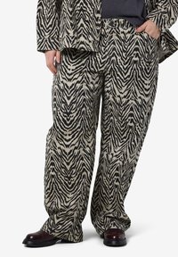 Pantalons beige amples avec un motif zèbre noir en zigzag, dotés d'une taille avec passants de ceinture, portés avec des bottines noires brillantes.