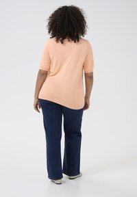 Licht perzikkleurig T-shirt met korte mouwen en een losse pasvorm, gecombineerd met donkerblauwe flare jeans en beige schoenen, gezien van achteren.