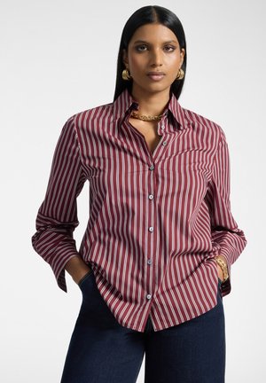 Mujer con el cabello largo y negro que lleva una camisa de botones a rayas rojas y blancas, pantalones oscuros y joyas doradas con las manos en los bolsillos.