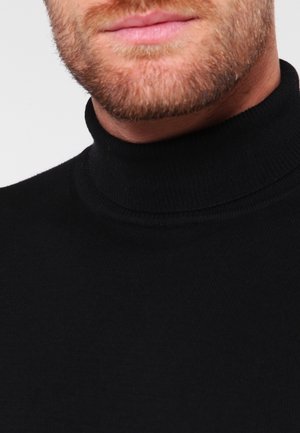 Uomo con la barba che indossa un maglione nero a collo alto, mostrando la parte inferiore del viso e il collo su uno sfondo uniforme.