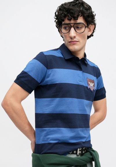 Πουκάμισο πόλο με μπλε ρίγες σε αποχρώσεις ναυτικού και ανοιχτού μπλε, κατασκευασμένο από βαμβάκι, με κοντά μανίκια και κεντημένο έμβλημα ασπίδας στο στήθος.