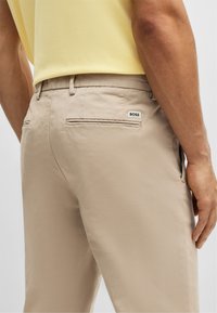 Beige trousers with a smooth texture, featuring two back pockets and a small black and white “BOSS” label on the right pocket.