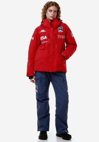 Giacca impermeabile rossa con colletto alto, cerniere e loghi; abbinata a pantaloni impermeabili blu scuro con tasche laterali e loghi.