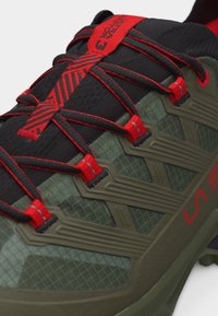Nahaufnahme eines grünen und schwarzen Wanderschuhs mit roten Schnürsenkeln und roten Akzenten, ausgestattet mit verstärkten Ösen und strukturiertem synthetischem Obermaterial.