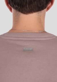 Svetlo roza pulover z rebrastim ovratnikom in majhno srebrno logotipno značko na hrbtni strani. Gladek tekstura s prilegajočo se zasnovo.