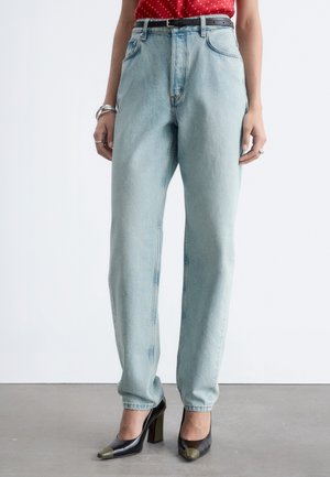 Donna che indossa jeans a vita alta azzurri chiaro, cintura nera, top rosso, scarpe con tacco alto nere con punte metalliche, in piedi su pavimento grigio.