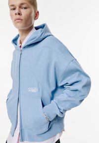 Haut en molleton bleu clair à zip en mélange de coton, arborant un logo sur le devant, des poches latérales et des poignets côtelés, avec une coupe décontractée et surdimensionnée.