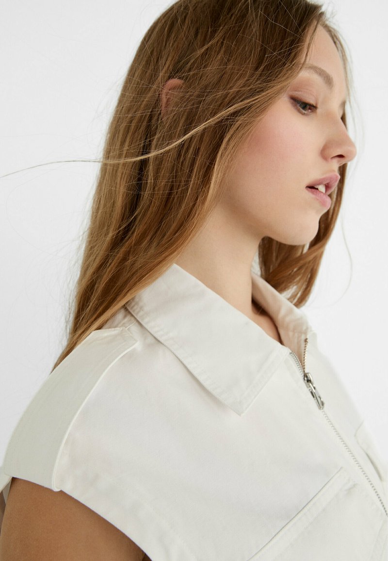 Chemise sans manches à col blanc, dotée d'une fermeture éclair et de poches avant. Tissu lisse avec une silhouette épurée et structurée.