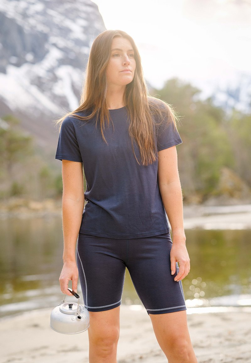 Marineblå t-shirt med korte ærmer og matchende cykelyder lavet af strækbart stof, holdt af en person udendørs med en sølvfarvet metalkedel.