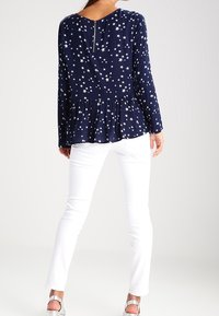 Maglietta a maniche lunghe blu navy con stampa a stelle bianche, stile peplum, dettaglio con zip sul retro. Indossata con pantaloni bianchi e scarpe con cinturino alla caviglia argentate.