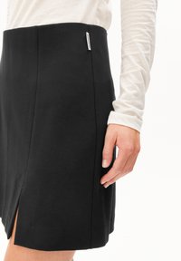 Frau in einem schwarzen Rock mit einem kleinen Seitenschlitz und einem weißen Langarmoberteil, die Hand entspannt neben dem Rock.