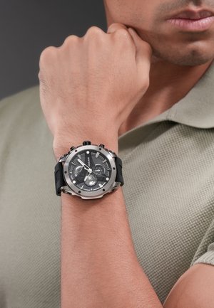 Man draagt een zilveren en zwarte chronograaf polshorloge met een zwarte wijzerplaat en meerdere subdials aan de linkerpols, en een groene polo met structuur.