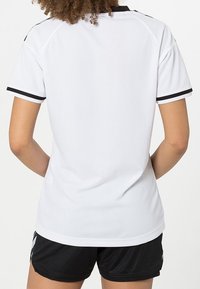 Persona con cabello rizado que lleva una camiseta deportiva de manga corta blanca con ribetes negros y pantalones cortos negros, de espaldas a la cámara.