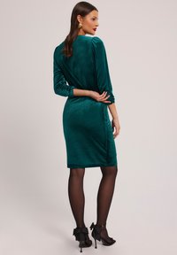 Turkusowa aksamitna sukienka z rękawami o długości trzech ćwierci, dopasowanym kroju i marszczeniami. Noszona z czarnymi szpilkami z dekoracyjnymi kokardami.