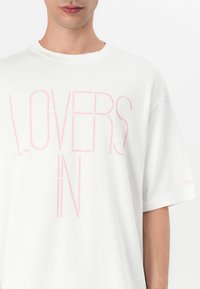 Bílý bavlněný tričko s výrazným vertikálním nápisem "LOVERS IN" v růžové barvě. Krátké rukávy a decentní branding na manžetě.
